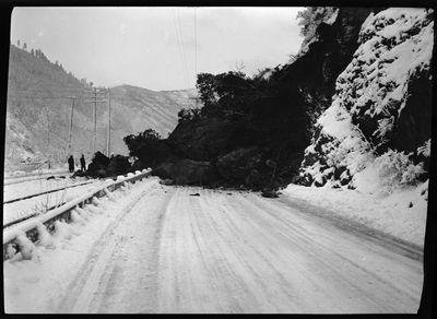Two people stand at the base of a landslide that fell into the roadway on the Wallace-Kellogg Highway. There is snow on the ground, but it does not appear to be an avalanche. Previously described as "Rock slide, Rocky Point, 1 mile west of Wallace, Idaho."