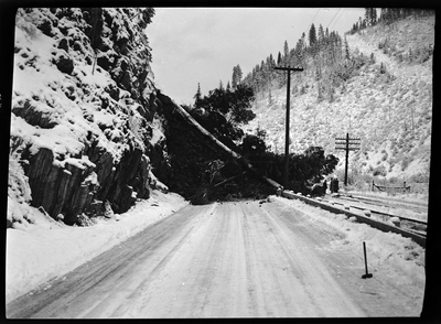 Several people stand at the base of a landslide that fell into the roadway on the Wallace-Kellogg Highway. Previously described as "Rock slide, Rocky Point, 1 mile west of Wallace, Idaho."