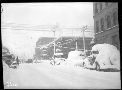 Photo of a street with several cars parked on either side of the road, most of which are covered in snow. There is a man standing in the street in the distance. It is actively snowing.