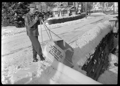 Photo of a man using a C. P. Carlson snow shovel to dump snow from the road. The shovel requires two hands to push it and holds more snow than the average snow shovel. There is a house behind the man. The side of the shovel reads: "Pats. Pend.; U.S. Ser. No. 738505; Can. Ser. No. 757075; C. P. Carlson; Duthie Idaho."