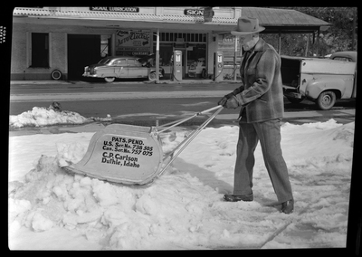 Photo of a man using a C. P. Carlson snow shovel to remove snow from the sidewalk. The shovel requires two hands to push it and holds more snow than the average snow shovel. The side of the shovel reads: "Pats. Pend.; U.S. Ser. No. 738505; Can. Ser. No. 757075; C. P. Carlson; Duthie Idaho." A road with cars on it can be seen behind the man.