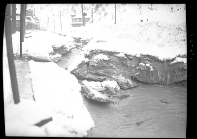 Photo of a creek running alongside some buildings in Wallace, Idaho, with snow surrounding the area. The buildings next to the creek have snow and icicles on them, as does the ground next to the creek. The water in the creek does not appear to be frozen over. Possibly from Pine Street or King Street.
