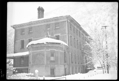 Photo of Our Lady of Lourdes Academy in Wallace, Idaho. There is a cross visible on part of the roof, so possibly a church or a religiously affiliated building. The surrounding ground, trees, and a bench are all covered in snow.