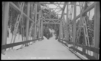 Photo of a snow covered bridge in a wooded area near Wallace, Idaho. The ground and trees are covered in snow.
