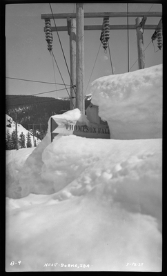 Photo of a wooden sign in the snow near Burke, Idaho. The snow comes up and mostly covers the sign. What is visible reads "Thompson Fall."