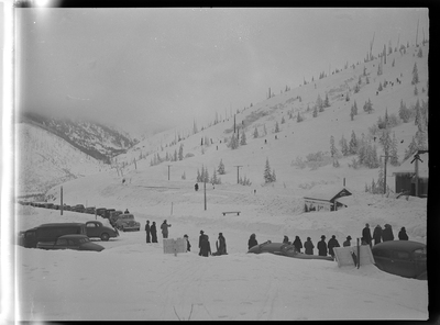 Photo of a group of people standing behind parked cars in the snow at Lookout Pass. There are a few buildings behind them, and snow covered trees are visible behind them.