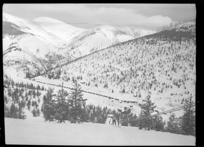 Photo of the ski lift at Lookout Pass. The ground is covered in snow, as are the trees, and a line of parked cars can be seen in the distance at the bottom of the hill.