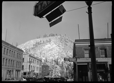 Photo of a hill covered in heavy snow and partially covered by trees. The upper part of a few buildings are visible.