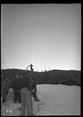 An unidentified person is mid jump at a ski jump event at Lookout Pass. There is snow on the ground and a crowd of people watching.