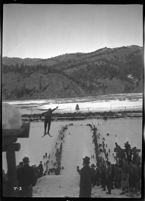 An unidentified person is mid jump at a ski jump event at Lookout Pass. There is snow on the ground and a crowd of people watching.
