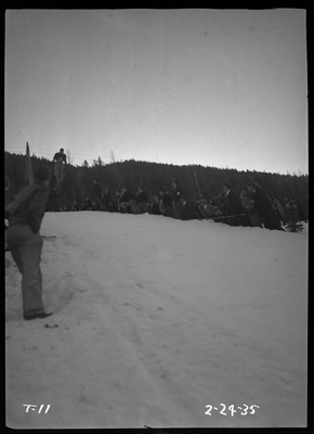 An unidentified person is mid jump at a ski jump event at Lookout Pass. There is snow on the ground and a crowd of people watching.