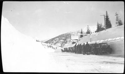 A line of cars are parked next to a packed wall of snow at Lookout Pass for a Ski Jump event. There is a group of people standing by the cars, but it is unclear what they are doing.