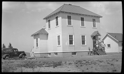 A group of unidentified people stand on the front steps of a house and appear to be posing for a photo. There a car parked next to the house with "J. W. Tabor" written on the side of it.