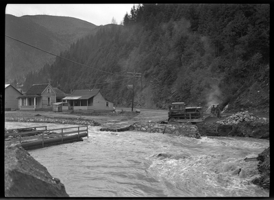 Photo of what appears to be a flood in a residential area, possibly near the Idaho Ski Club Lodge building. The water reaches up to the road across the water where there are several houses and some machinery.