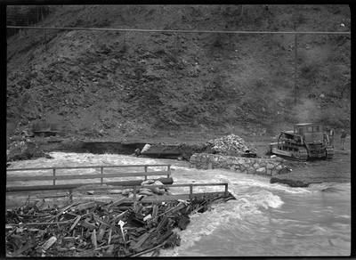 Photo of what appears to be a flood in a residential area, possibly near the Idaho Ski Club Lodge building. The water reaches up to the road across the water where there is some type of machinery.