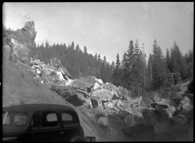The aftermath of the blast. The dust has settled, and large chunks of rock are visible, piling at the bottom of the hill. There is a car in the foreground.
