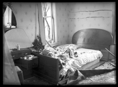 Damage done to a bedroom in N. J. Osborne's house by the Sunshine Mine powderhouse explosion. The walls and window are damaged, and there is debris all over the room and bed.