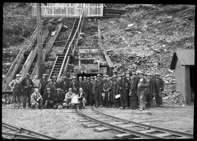 Hecla Mining Company miners posing together for a photo with their recently granted safety award. They are all standing outside of the mine entrance that has two signs posted. One reads, "Safety First," and the other reads, "No Smoking on coaches going in or out of tunnel."