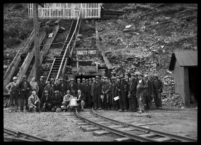 Hecla Mining Company miners posing together for a photo with their recently granted safety award. They are all standing outside of the mine entrance that has two signs posted. One reads, "Safety First," and the other reads, "No Smoking on coaches going in or out of tunnel."
