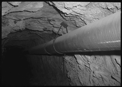 Image of a long pipe that is hung up underground at Bunker Hill. The pipe is held up by chains attached to the rocky ceiling and walls.