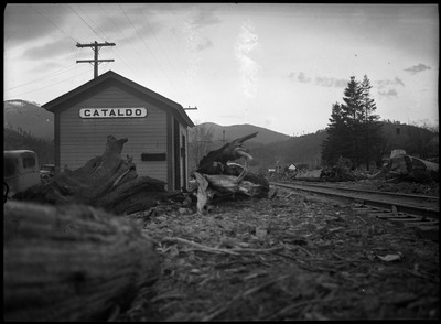 Downed trees and other plant debris litter a train track after the Wallace flood. Several buildings and telephone poles are visible in the background, and the closest sign reads "Cataldo."