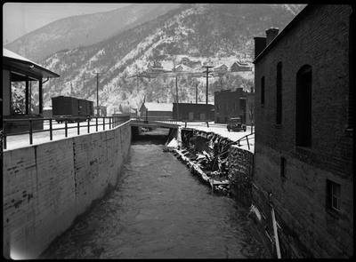 Debris falls from the side of the road into a nearby river during the Wallace flood. A few buildings, rail cars, telephone poles, cars, and a bridge are in the background.