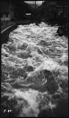 Water rushing towards a bridge and buildings. A person can be seen on the street watching the water.