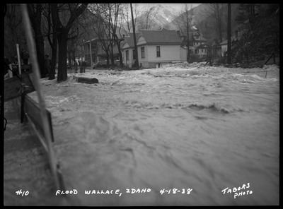 Water flowing in the street during a flood. People are standing overseeing the water on the left. Standing buildings are nearby the floodwaters.