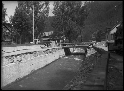 Image of a bridge with water running beneath it. A man is walking on the bridge as two others stand on the left end of the bridge. The two men stand next to a truck. A boy with his bicycle is on the right. Piles of debris are both ends of the bridge. A few buildings are also in view.