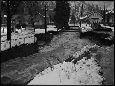 Water rushing through Wallace, Idaho during a flood. A few people are walking in the distance on the right. There is snow on the ground and on surrounding buildings.