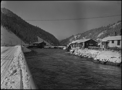 View of water during a flood. It appears to have eroded the area near some buildings covered in snow. One building has sustained damage and is falling into the water.