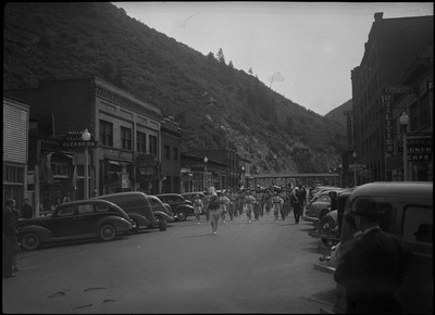 Band members, a few women and mostly men, in the Benevolent and Protective Order of Elks parade. Spectators are watching near the cars parked on the street.