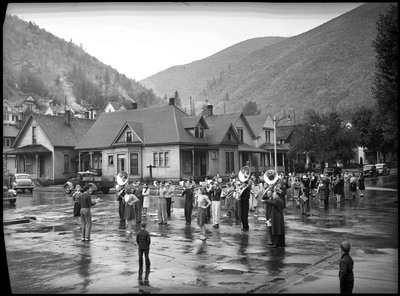 Band playing in the Defense Bond Parade. The ground is wet, probably from rain. Two children watch from the sidewalk.