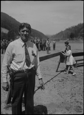 A man, possibly a police officer, poses with a cigarette in his hand and a gun attached to his belt. The Co. K. Kellogg home guards stand in the distance. Two children watch behind a fence.