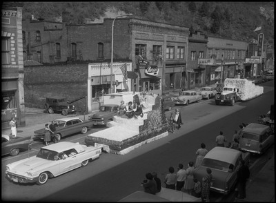 Four women sitting on a float during a Benevolent and Protective Order of Elks roundup parade. Another float follows behind this one. Spectators watch by parked cars along the side of the street.