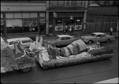Three women sitting on a very decorated float. There is text at the front of the float. Spectators watch from a car and from the sidewalk.