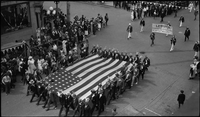 Men wearing suits and hats with fake antlers hold a large flag during a Benevolent and Protective Order of the Elks parade. Several men follow behind and hold a sign that reads, "LEWISTON The best damn Town in the whole damn World (AFTER WALLACE)" Crowds of spectators are watching the procession.