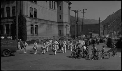 A marching band and crowds of children wait in the street by a large building for the back-to-school parade. Several children hold signs. A few children are on their bikes on the right.