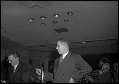 A man stands near a microphone at the March of Dimes even at the B.P.O.E. temple and reading or singing from a paper in his hand. Part of a band can be seen playing in the background and another man on the left.