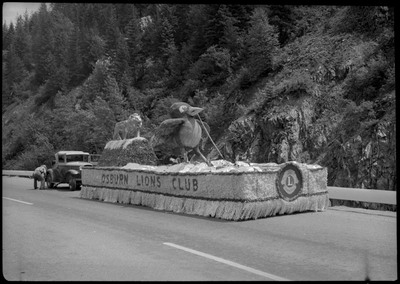 The Osburn Lions club float decorated with a big bird and lion during the Silver Jubilee. A man is crouched beside a car behind the float.