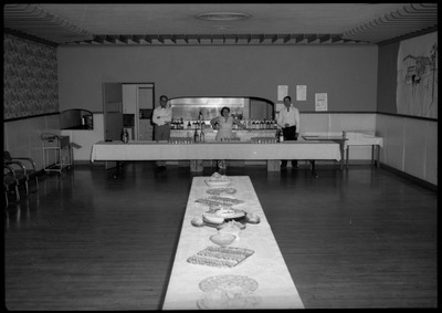 A table with food in the middle of a room as three people stand in the back of the room near what looks like a bar area.