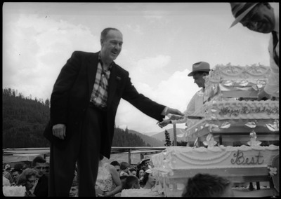 A man cutting a huge cake during the Silver Jubilee with a smaller knife. Other people are also standing and watching near the cake.
