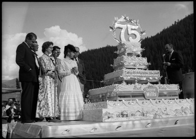 People standing on a stage next to a large cake during the Silver Jubilee. A man speaks into a microphone on the right. The cake is decorated with the number "75" and the text, "Happy Birthday Wallace, Silver Jubilee" "Best Wishes from General Mills."