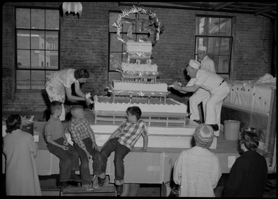 Two people use frosting bags to decorate the Silver Jubilee cake. The cake is on the flatbed of a Builders Hardware Co. truck. Three children and one adult are on the flatbed as three other people watch on the ground.