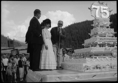 Five people on a stage with a large Silver Jubilee cake. One of the men stands near the microphone. Other people can be seen standing on the ground.