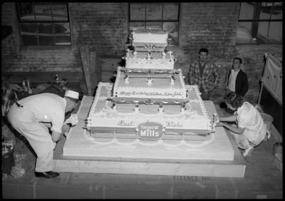 Two people use frosting bags to decorate the Silver Jubilee cake. Two people are watching.