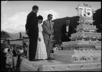 People standing on a stage next to the Silver Jubilee cake. Other people are watching on the ground.