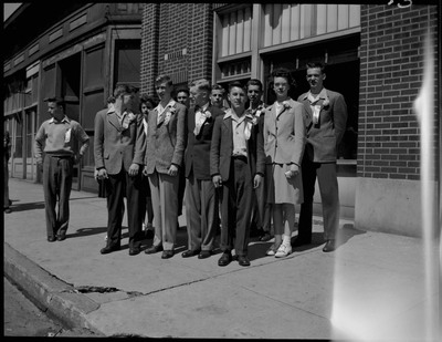 A group of teenagers with ribbons pinned to their shirts during Boys and Girls Week. The teenagers are standing on the sidewalk in front of a brick building.