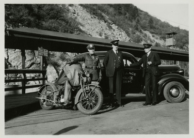 Possibly Wallace police officers in uniform standing near a motorcycle and a Wallace Police Department car outside. Image previously described as: "Tom Barrett, Vic Langley, Charles Pugh. Police photographs."