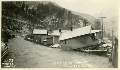 A river encroaches on several buildings during the Wallace flood. A least one of them appears to have sustained structural damage and appears to be falling into the river. There is snow on the ground and a bridge nearby. There is a person and several telephone poles in the background.